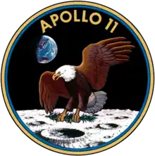 Circular insignia: eagle with wings outstretched holds olive branch on Moon with Earth in background, in blue and gold border.