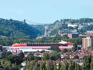 In the foreground twentieth century housing can be seen amidst trees and on the right a tower block of flats. In the middle distance a complex of red coloured buildings can be seen and behind that a steep sided gorge with a suspension bridge spanning it. Eighteenth century terraces on the right side of the gorge, the slopes of which are heavily wooded and a tower can be seen in the distance on the skyline.