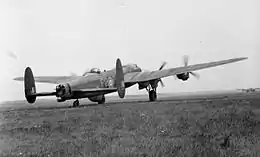 Rear three-quarter view of four-engined military aircraft with twin tailfins, parked on landing ground