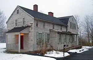 A two-story gray wooden house with light green shutters seen in winter, with snow on the ground. A long wing with two brick chimneys projects towards the left from a higher rear section. At lower right is a black mailbox with "5Ave" on it next to a clear paved driveway.