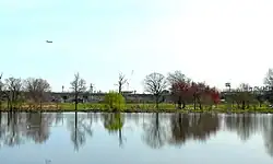 Meadow Lake with baseball fields behind it.  Interstate 95 borders the park, with the Philadelphia Naval Shipyard behind it. Above an airplane begins its landing at Philadelphia International Airport.