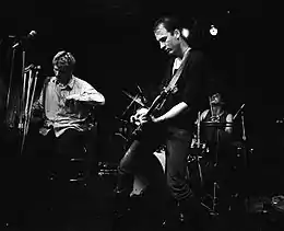 The band Flipper is performing at a club. From left to right are the singer, drummer, and electric guitarist. The singer is seated on a stool, and he is holding a pair of crutches.