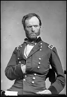 A black-and-white photograph of a man seated with his left hand inside of his jacket. His body is facing the right diagonal while his head is looking to the left.