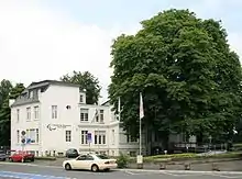 A white building with trees next to it fronting a street with a car driving past