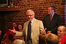 Barr stands in front of a brick wall in a vanilla suit, with his arms stretched to convey a point, before the small crowd around him