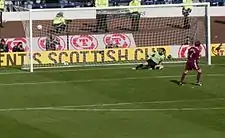 A football player scores a goal against the opposing goalkeeper from a penalty-kick. Stewards and camera-operators are visible behind the goal net.