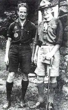 two men outdoors, both wearing Scout uniforms: short sleeve shirt, shorts and knee socks, the older man on the right wearing a campaign hat and holding a cane