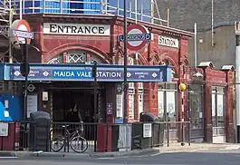 A red-bricked building with a rectangular, white sign reading "ENTRANCE" in black letters and a rectangular, blue sign reading "MAIDA VALE STATION"