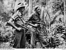 Two men wearing military style uniforms, one wearing a slouch hat, the other a general service cap, carrying weapons in a jungle setting