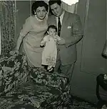 A woman and man, both in their thirties and both dressed in 'Sunday best', hold a similarly dressed very young girl standing on the arm of a floral-print sofa.