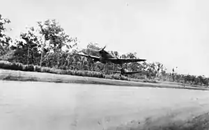 Two single engined propeller-driven monoplanes fly just above a cleared area. A thick forest is visible behind the aircraft.